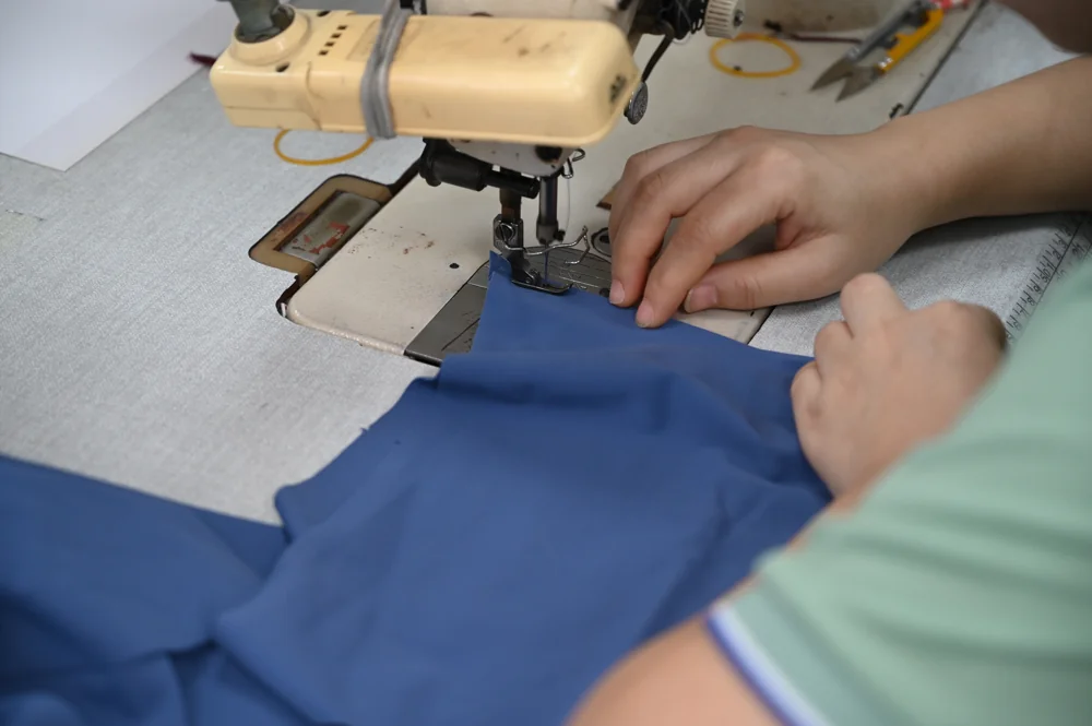 Hands feeding blue fabric under the needle of a sewing machine in the sample room