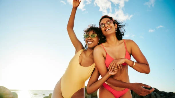 Two young women wearing yellow and pink swimwear laughing happily on a sunny beach with ocean and blue sky background